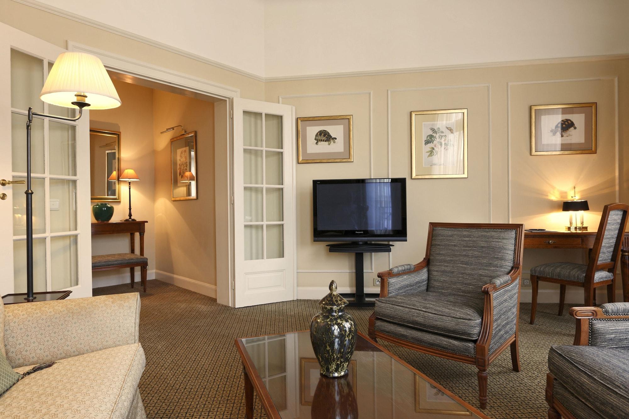 Hotel Le Plaza Bruselas Habitación foto The image shows a cozy and elegantly designed living room area. There is a comfortable sofa and armchairs arranged around a coffee table. A large television is mounted on a stand, facing the seating area. The walls are adorned with framed artwork, an
