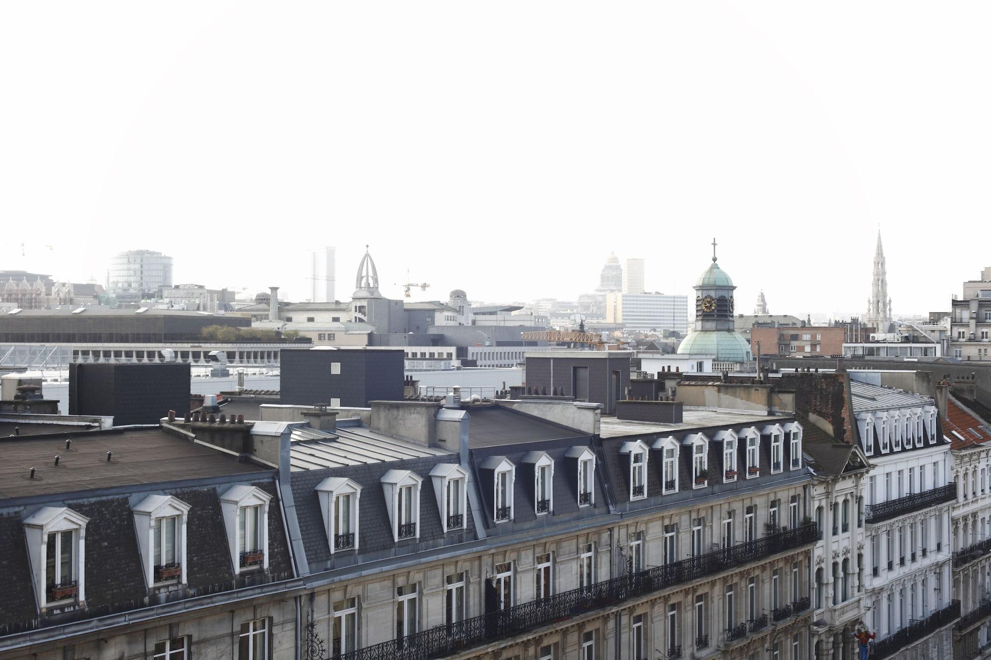 Hotel Le Plaza Bruselas Exterior foto The photo depicts a skyline view of a city, featuring various rooftops with distinctive architectural styles. You can see multiple buildings, including some with sloped roofs and dormer windows. In the background, there are taller structures, possibl