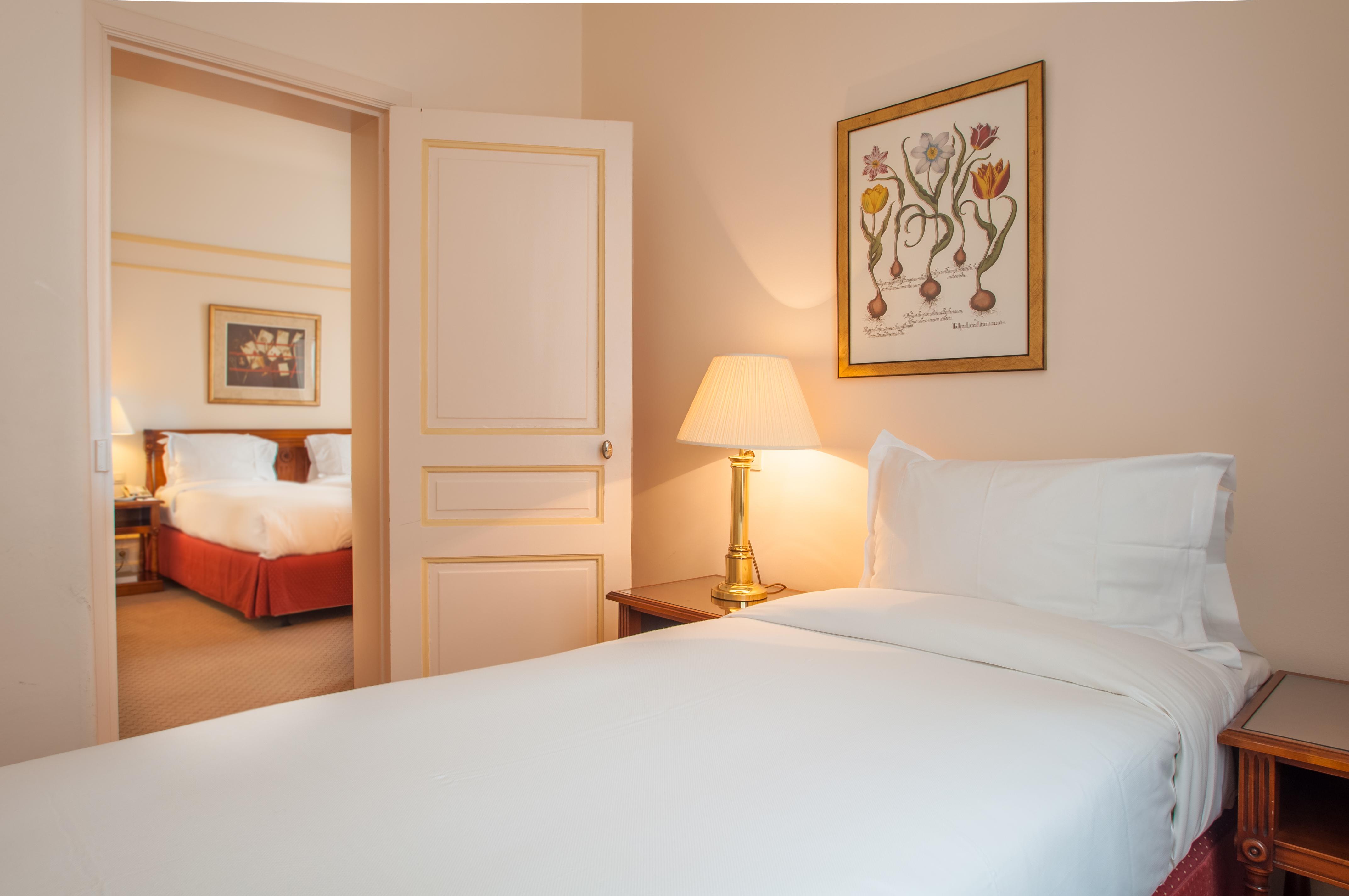 Hotel Le Plaza Bruselas Exterior foto The photo shows a cozy hotel room with a single bed covered in white linens. There's a bedside table with a lamp providing soft light. On the wall, there is a framed piece of artwork featuring flowers. In the background, through an open door, a secon