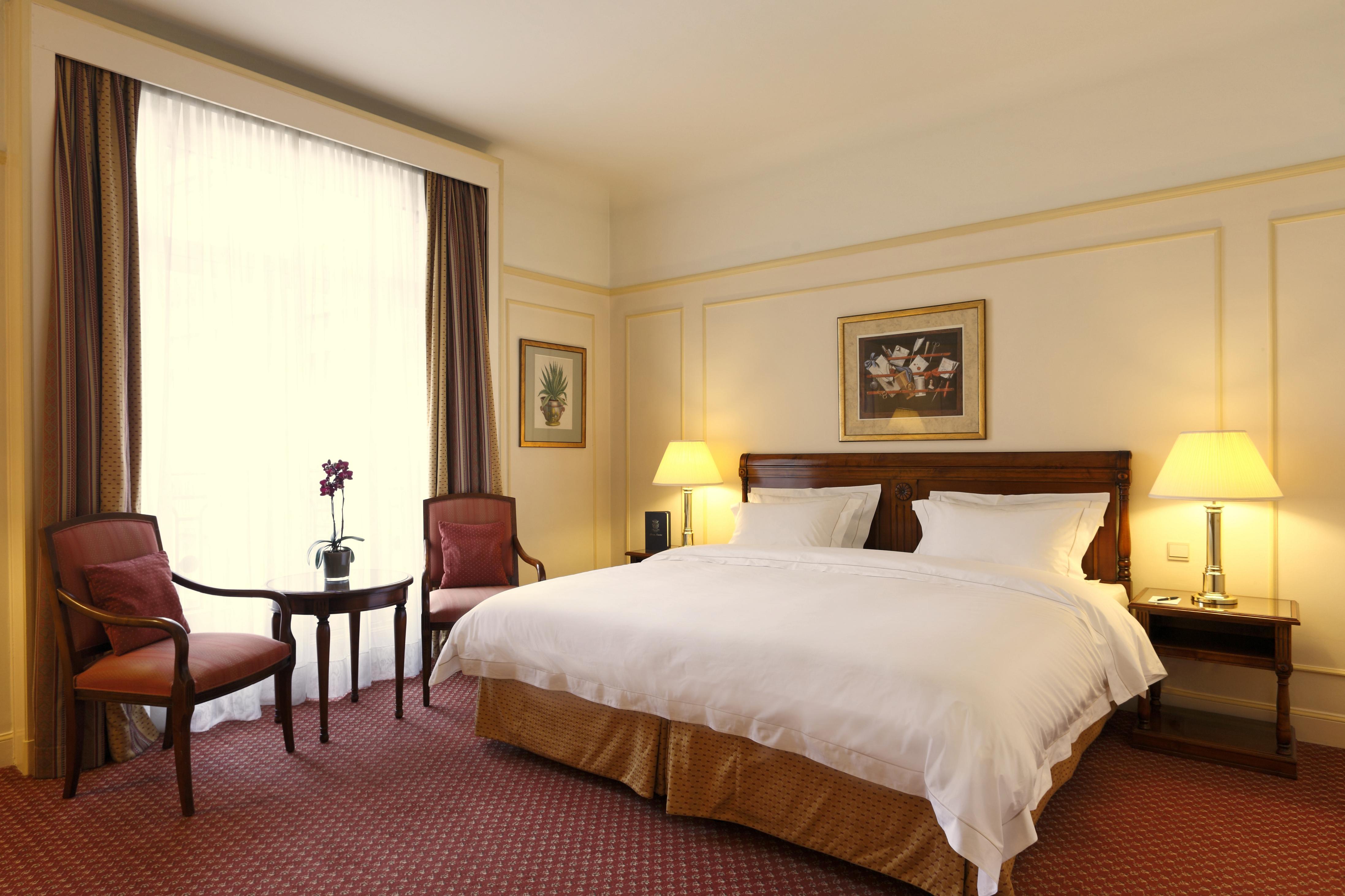 Hotel Le Plaza Bruselas Exterior foto The photo shows a cozy hotel room featuring a double bed with white bedding and a gold accent at the base. To the sides of the bed, there are two bedside lamps providing soft lighting. There are two upholstered chairs and a small round table position
