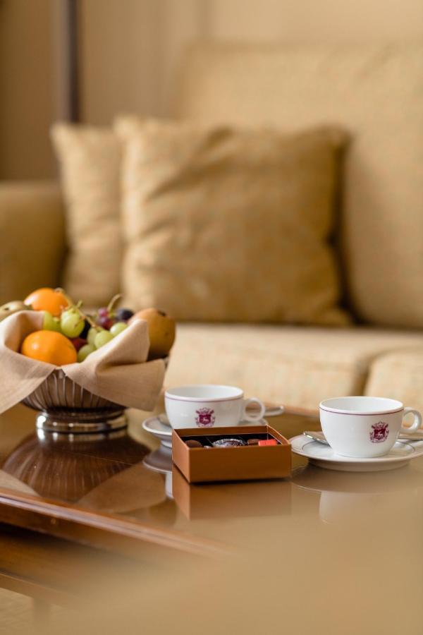 Hotel Le Plaza Bruselas Exterior foto The photo shows a cozy setting with two small white cups placed on saucers, likely for coffee or tea. Next to the cups, there is a small box of chocolates. In the foreground, a silver bowl holds a variety of fruits, including oranges, grapes, and oth