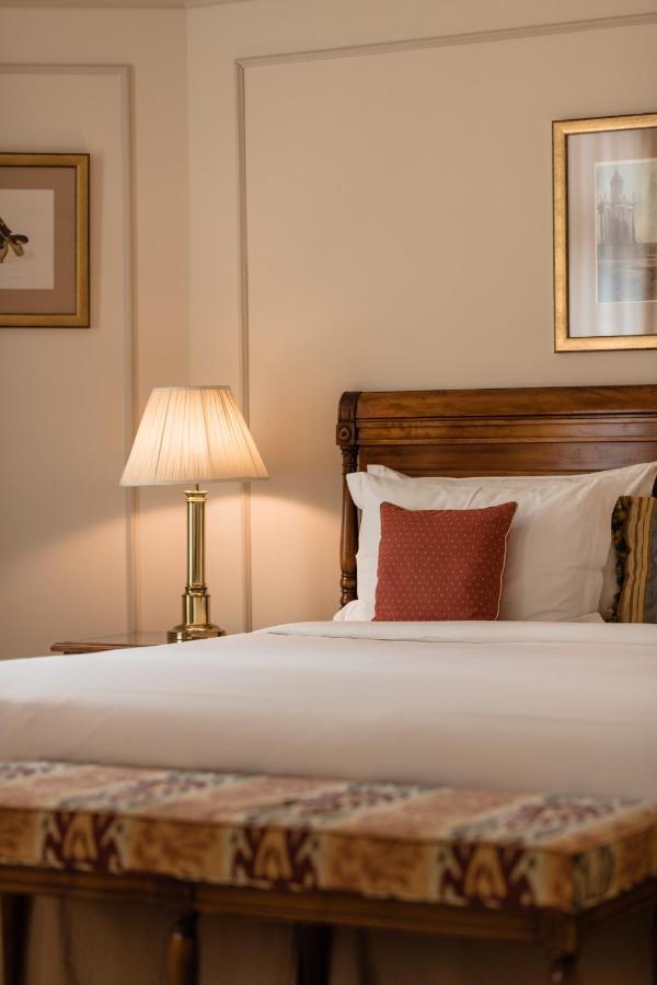 Hotel Le Plaza Bruselas Exterior foto The photo shows a cozy bedroom setting. There is a wooden bed with white bedding and a decorative red pillow. Next to the bed, there is a bedside lamp with a cream-colored lampshade. The walls are adorned with subtle decorations, likely framed artwor