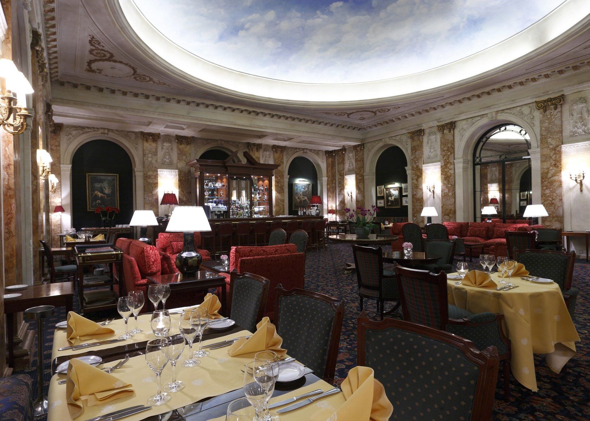 Hotel Le Plaza Bruselas Restaurante foto The image depicts a luxurious restaurant interior. It features elegantly arranged tables with yellow tablecloths and napkins. The seating area includes plush red couches and dark wooden chairs. The walls are adorned with ornate decorations, and there