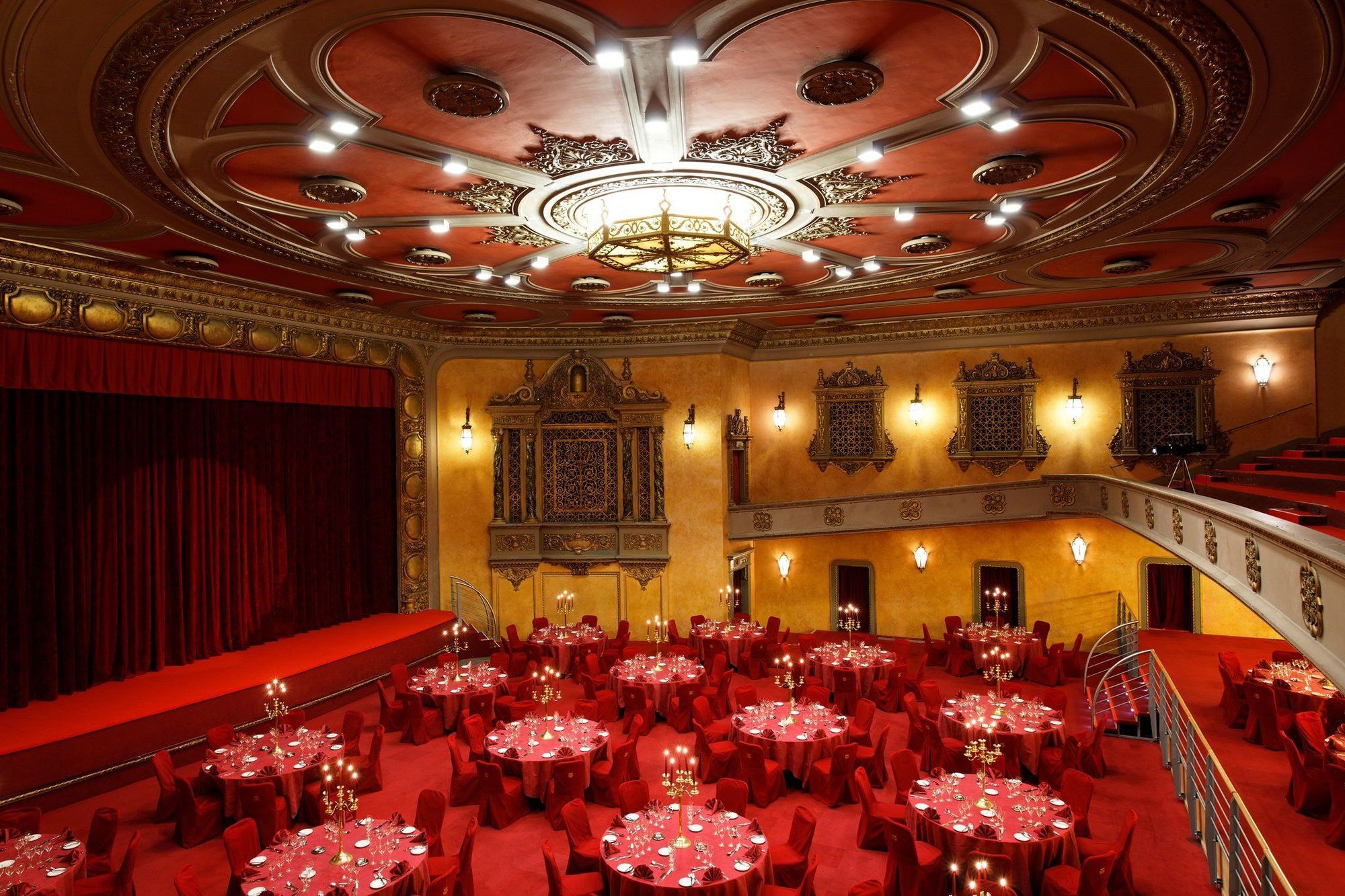 Hotel Le Plaza Bruselas Restaurante foto The photo shows an elegant theater interior, featuring a stage with red curtains and a beautifully designed ceiling with intricate details and lighting fixtures. Below the stage, there are round tables set with red tablecloths and candles, arranged i