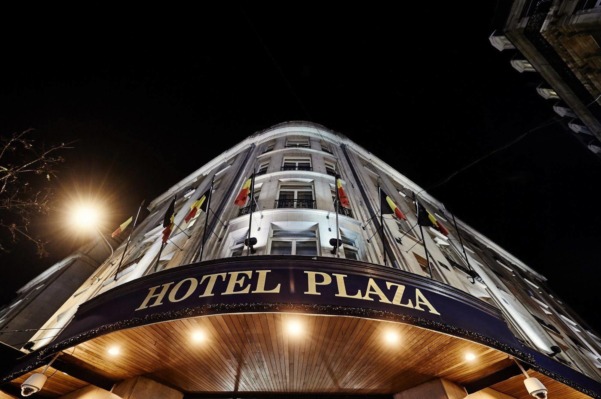 Hotel Le Plaza Bruselas Exterior foto The photo shows the exterior of a hotel called "Hotel Plaza." It is taken at night, and the hotel has a prominent entrance that features a curved facade. The building is illuminated by exterior lights, highlighting its architecture. There are flags d